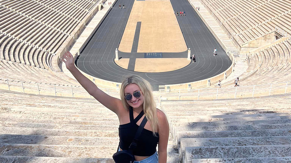 Student poses with one hand raised in front of ancient stadium track.