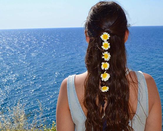 Woman with long brown hair decorated with chain of six daisies looks out over ocean.