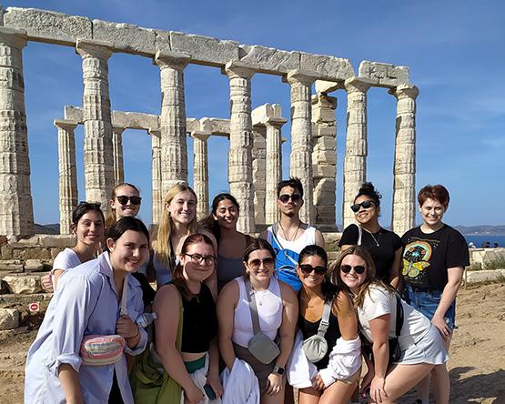 Two rows of students pose in front of ancient marble columns of a temple.