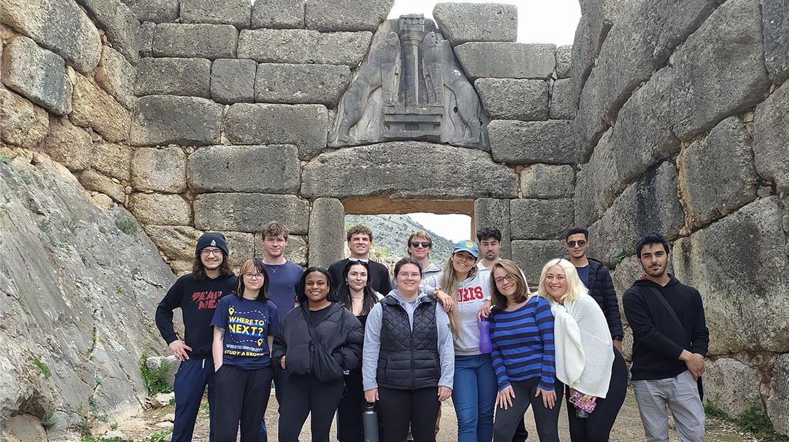 Group of Webster students pose in front of Lion's Gate in Mycenae, which has a carved stone piece over doorway with two lions.