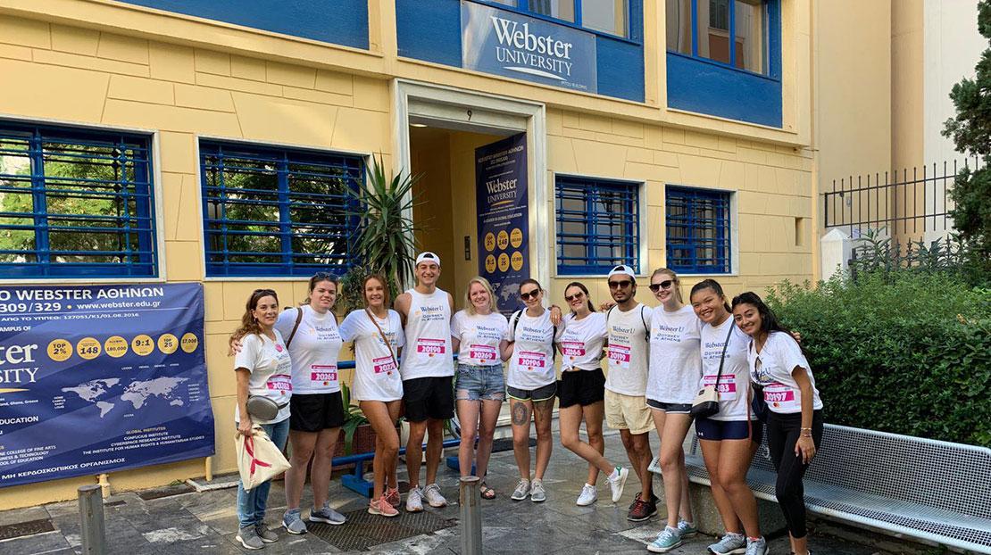 Line of Webster students dressed with runner numbers pose in front of the Athens campus building.