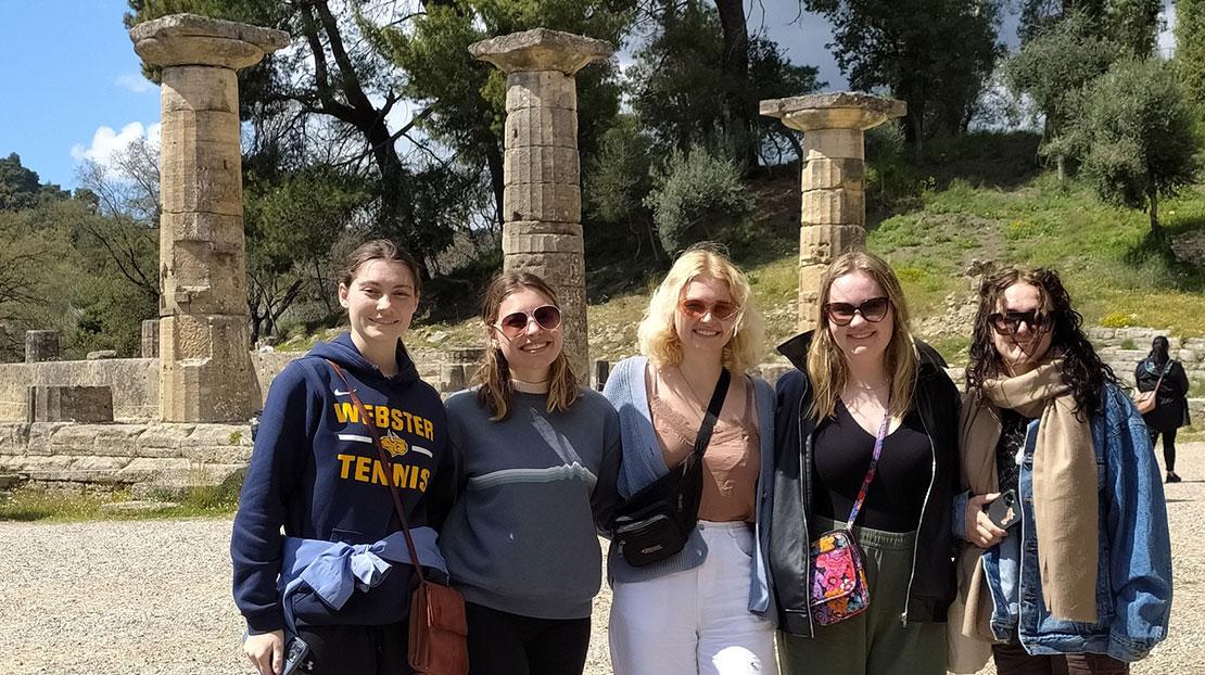 Five students wearing warm clothing stand in front of three pillars in Delphi.