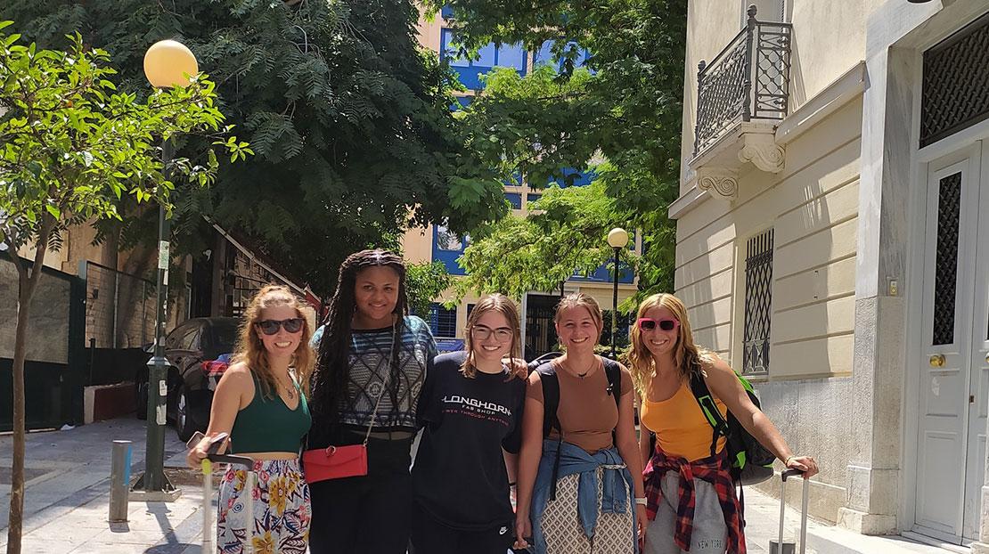 Five students pose with suitcases on street in front of Athens campus building.