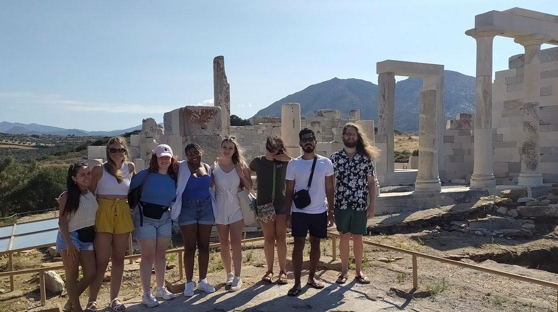 Eight students pose together at the Temple of Demeter in Naxos, with ancient marble columns behind them.